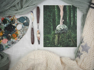 Fine art print of barefoot ballerina legs standing demi-pointe atop a shattered mirrorball in a forest. Set next to two palette knives, a paint-covered palette, and a Taylor Swift cardigan.