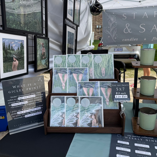 A booth display at an outdoor market or craft fair featuring art prints, a sign reading 'What is a Giclee Print?', candles in small pots, and a backdrop banner for 'Starlight & Sage Co.'