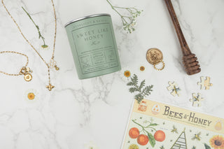 Green candle labeled 'Sweet Like Honey' with a honey dipper, necklace, and decorative items on a marble surface.