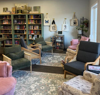 A cozy, well-lit reading area inside Flutters & Flowers Bookshop featuring multiple upholstered armchairs and armchairs with wooden frames, a large bookshelf filled with books, a floral area rug, and a white paneled ceiling.