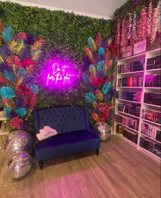 A decorated corner inside Sweeter Than Fiction Bookshop in Charleston, SC. A green foliage wall, colorful artificial palm leaves, a glowing pink neon sign reading 'Do it for the pot', a dark blue velvet tufted sofa with a pink heart pillow, mirrored disco balls, and white shelving filled with pink and purple items.